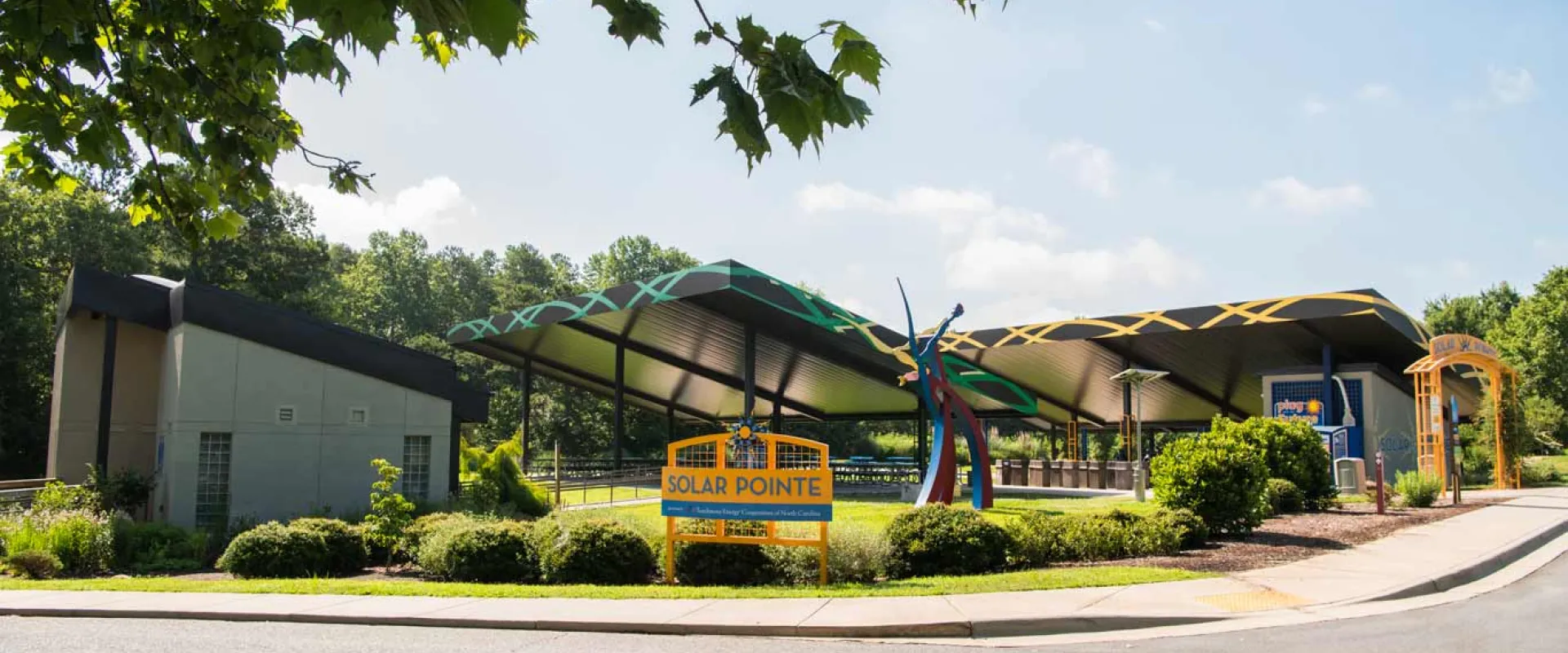 Two large, metal awnings that are covering a paved area that is sprinkled with picnic tables, stand next to a small, brown building. There are yellow signs in front of them with the text "Solar Pointe" which are posted in a curated garden bed.