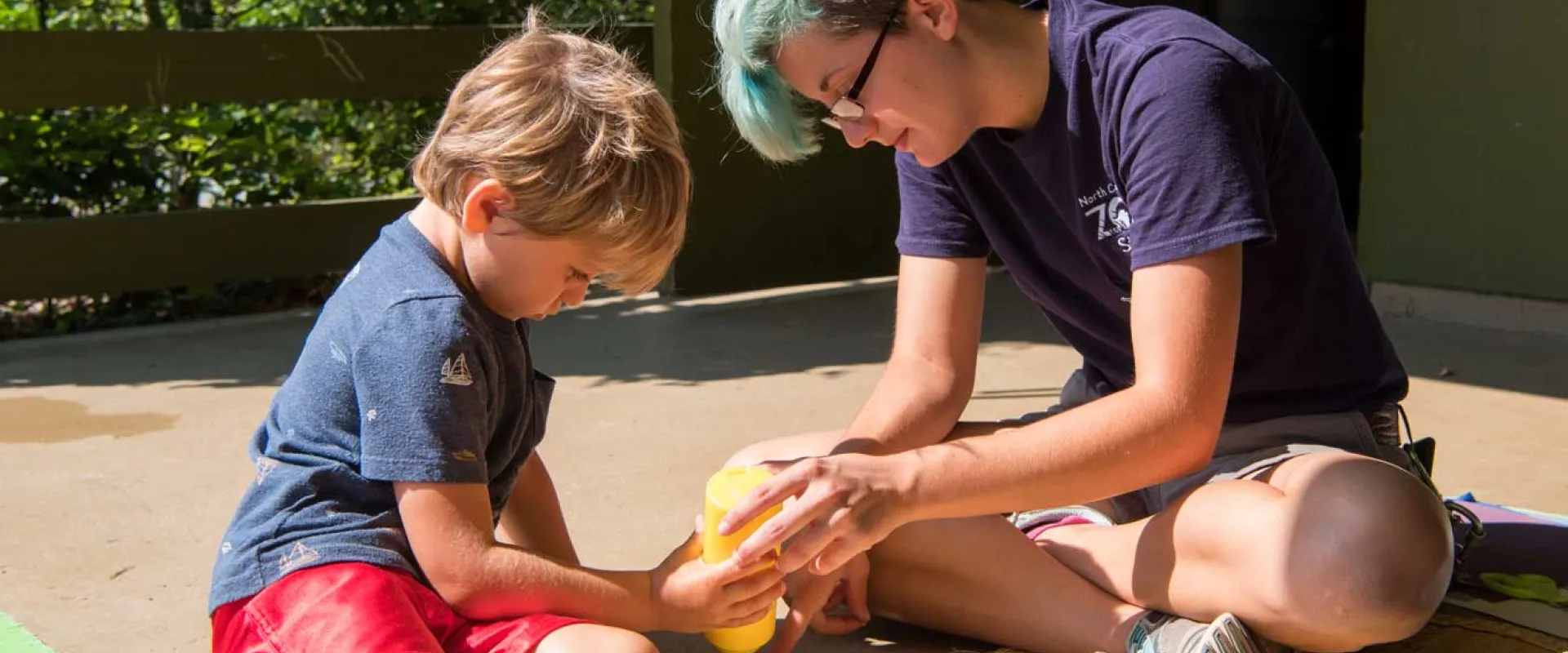 An adult with short blue hair sits on the ground next to a child. They are holding a bottle of what appears to be glue and seem to be working on a craft together on a covered porch area.