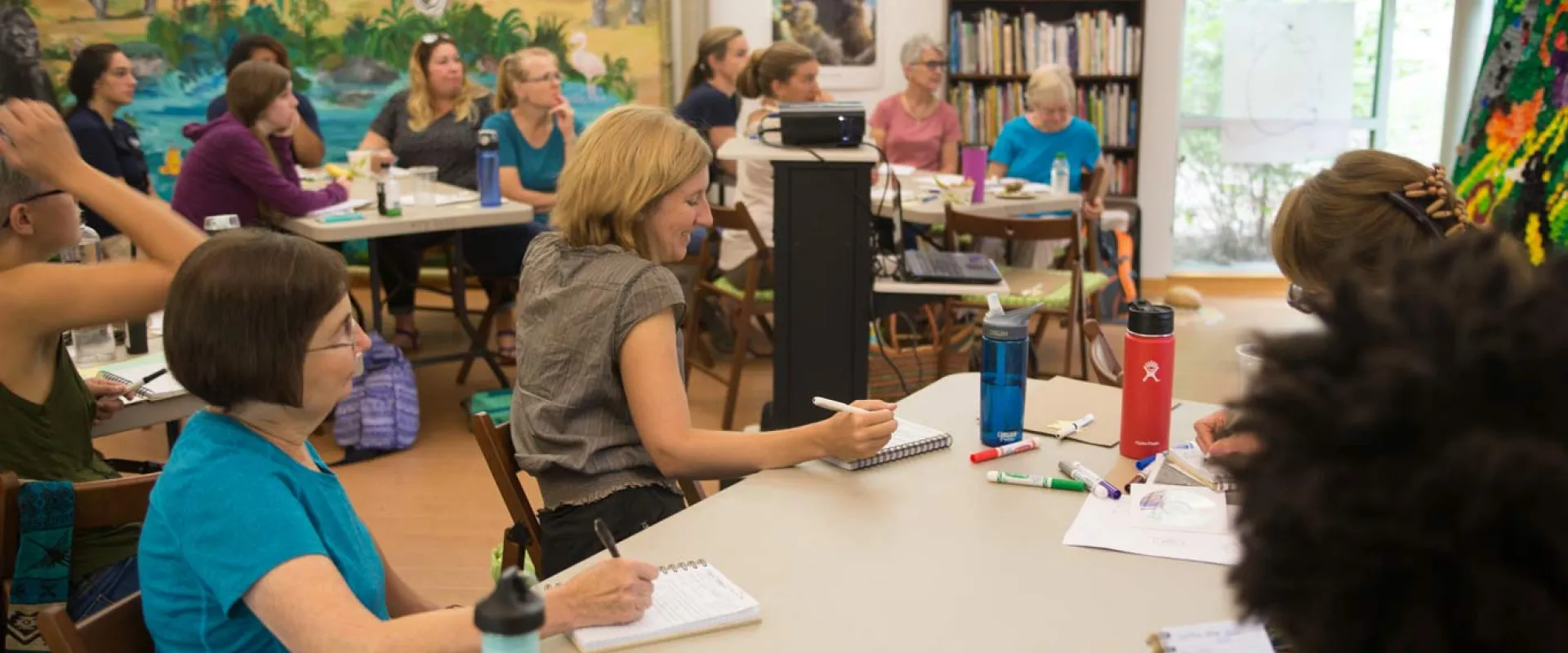 A group of about fifteen adults, mostly women, are seated at light-colored tables in a bright room, facing a woman in a blue shirt who is standing and gesturing towards a projected image on a white board. Colorful animal artworks, including a large polar bear and a macaw, adorn the walls behind the standing woman.