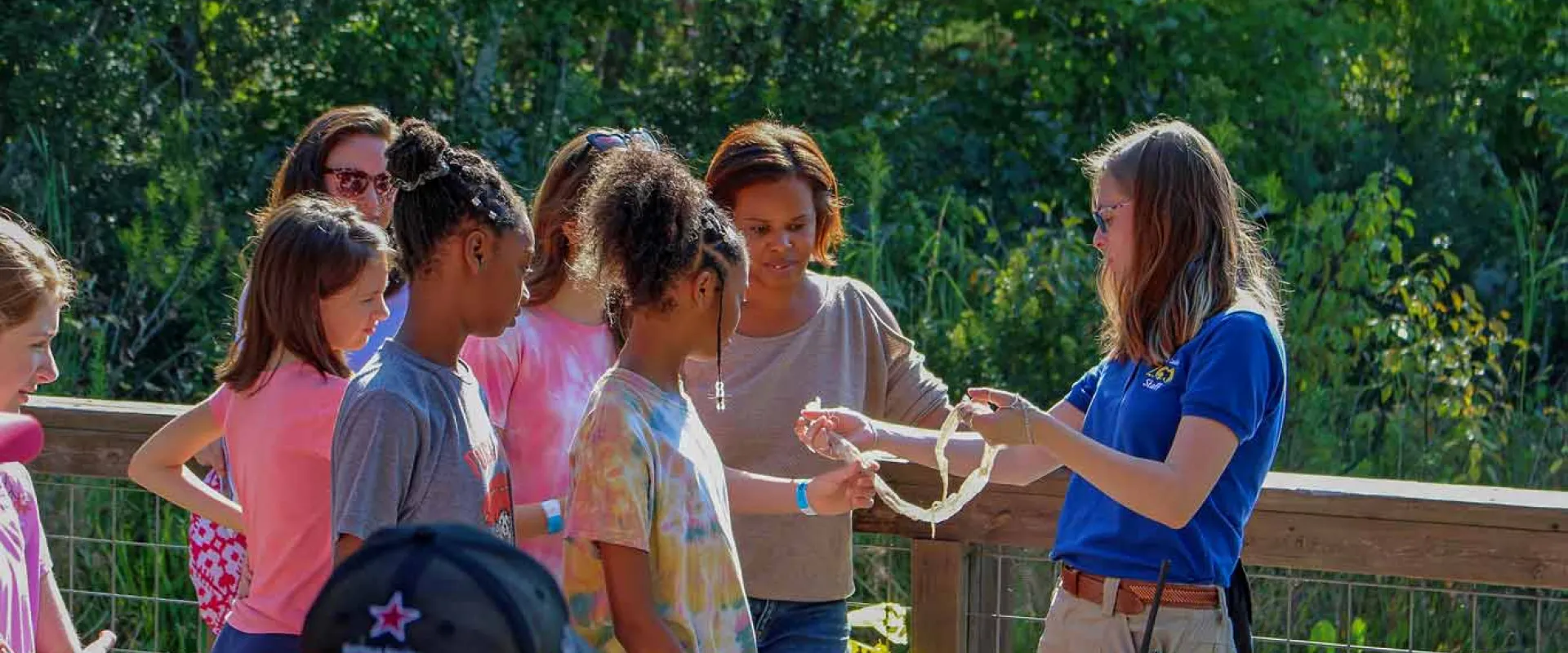 A woman with brown hair wearing a NC Zoo uniform stands smiling in front of a fence holding what appears to be a snake skin. She is showing a group of children who are standing in front of her. They appear to be standing on a wooden deck, or bridge, with dense green foliage behind them.