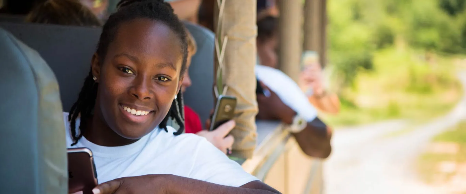A group of kids sit in large bus seats on the back of an open air safari truck. The focus is on a young person with shoulder length dark braids, who is staring at the viewer, smiling while holding a cellphone in one hand and leaning an elbow out of the side of the truck.