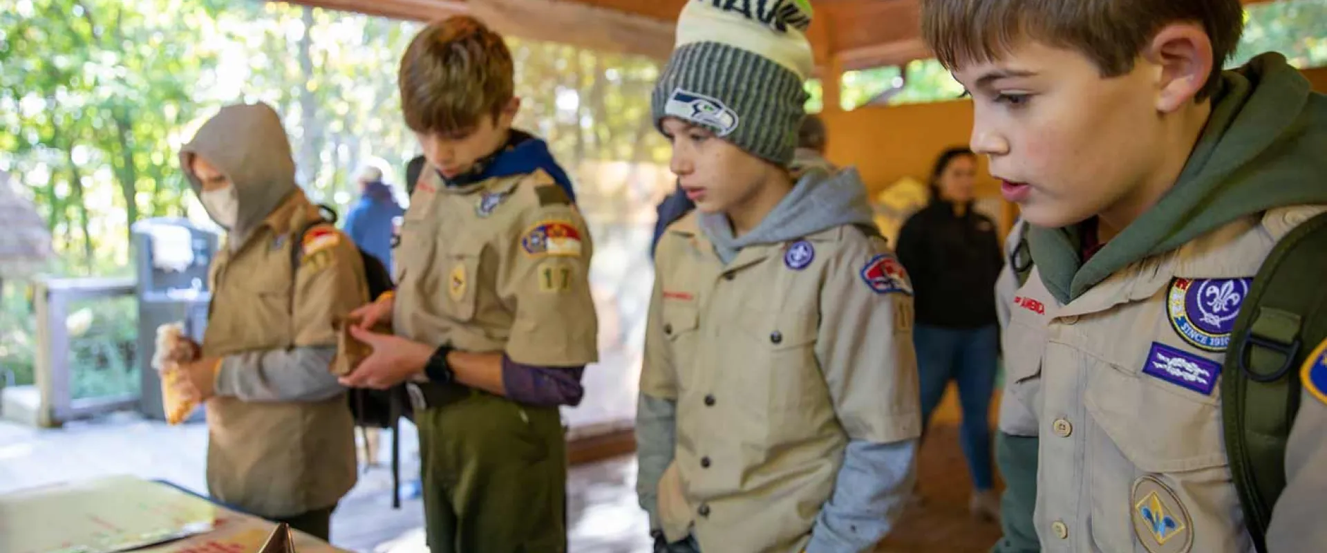 Four young people appear to be gathered around a table outdoors. They are wearing tan boy scout uniforms that are covered with patches and appear to be listening to another individual who is not visible to the viewer. On the table are various objects, including what appear to be drawings and graphs. The setting looks like a covered outdoor area with stone pillars or walls in the background.