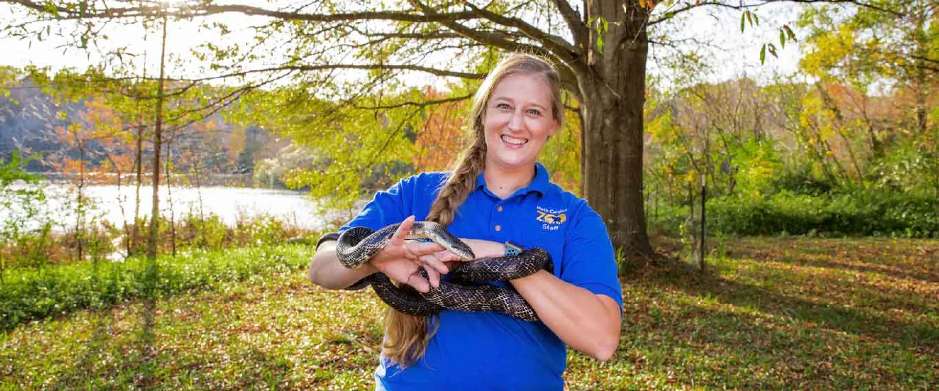 A woman with a long, blonde braid, wearing a blue collared shirt is standing next to a lake in the woods. She is smiling at the camera and holding a very long, black snake in both hands as it wraps around her hands and forearms.