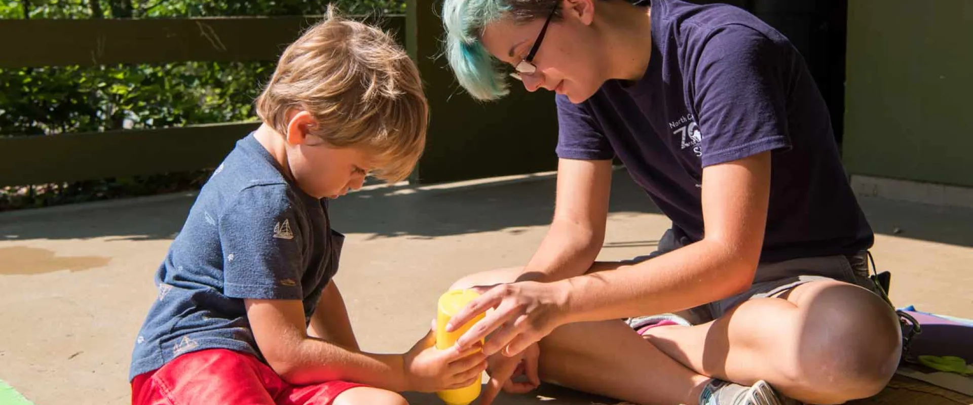 An adult with short blue hair sits on the ground next to a child. They are holding a bottle of what appears to be glue and seem to be working on a craft together on a covered porch area.