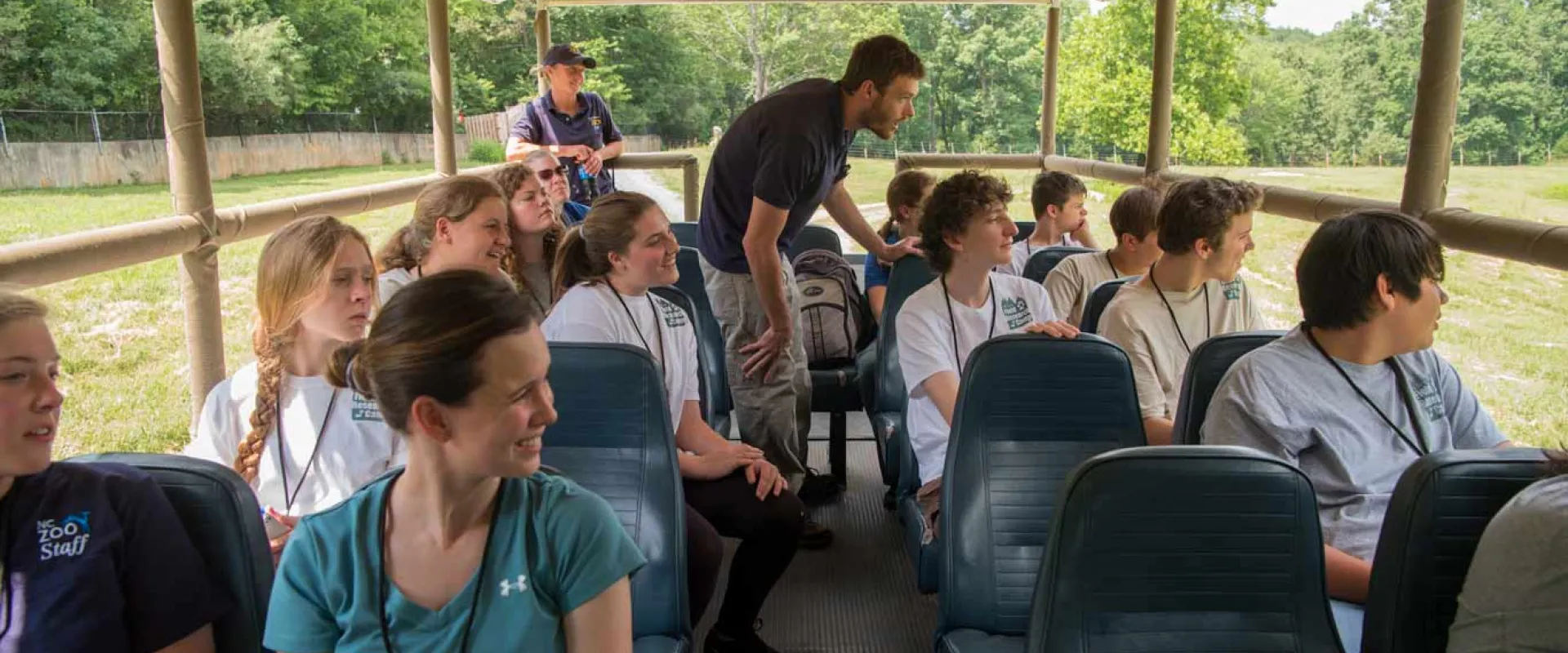 A group of students viewing animals from the open-air Zoofari bus as part of their Wild Science Camp experience. It is driving through a large, open field and trees are visible in the background.
