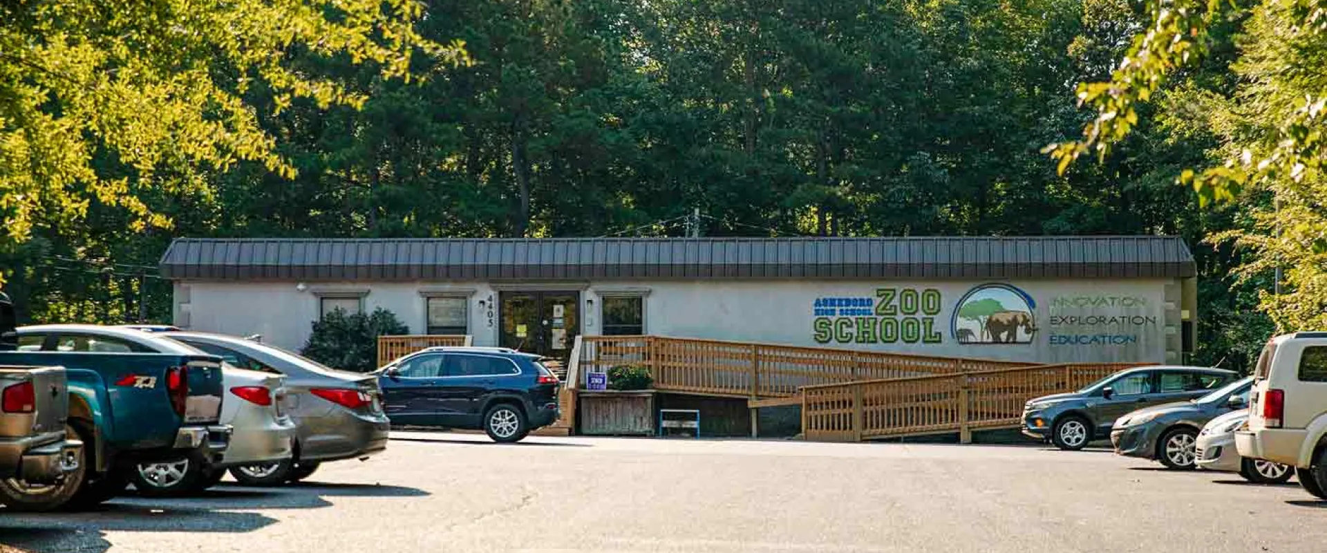 A wide angle shot of a long, grey trailer building with green text that says "Zoo School" on the front right. There is a long wooden ramp that winds from the front door to the parking lot which is filled with cars. There are trees all around the building and lot.