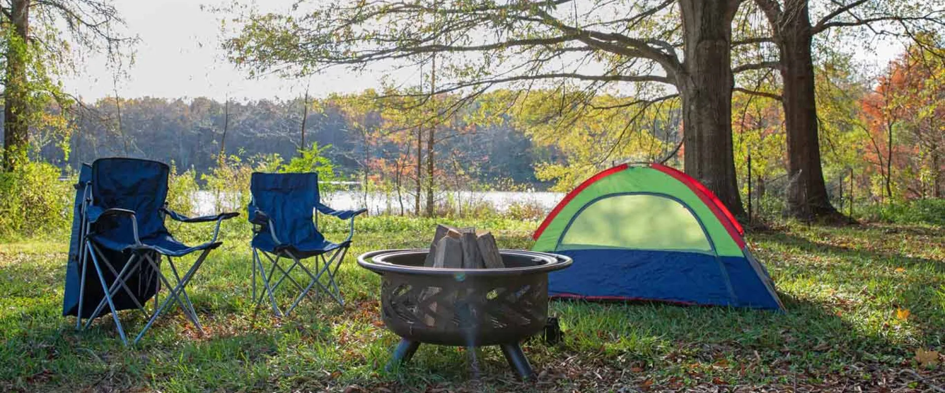 A serene campsite consisting of a small green, red and blue tent, a fire pit and two camping chairs that has been set up amidst the trees, next to a calm lake.