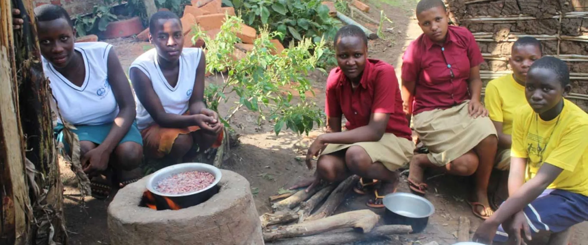 Six young women or girls are crouching around a traditional outdoor cooking fire set up on the ground, likely in a rural African community. In the center, a cooking pot filled with red beans sits on a raised clay or stone stove. The girls are looking towards the viewer, wearing a mix of school uniforms (white vests and skirts/wraps, or maroon shirts) and yellow t-shirts. More cooking pots and wood are visible on the ground.