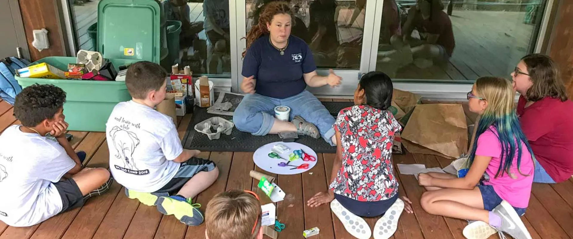 A teacher is leading a group of six children sitting on a wooden porch, demonstrating a craft project using repurposed materials like cardboard and egg cartons.
