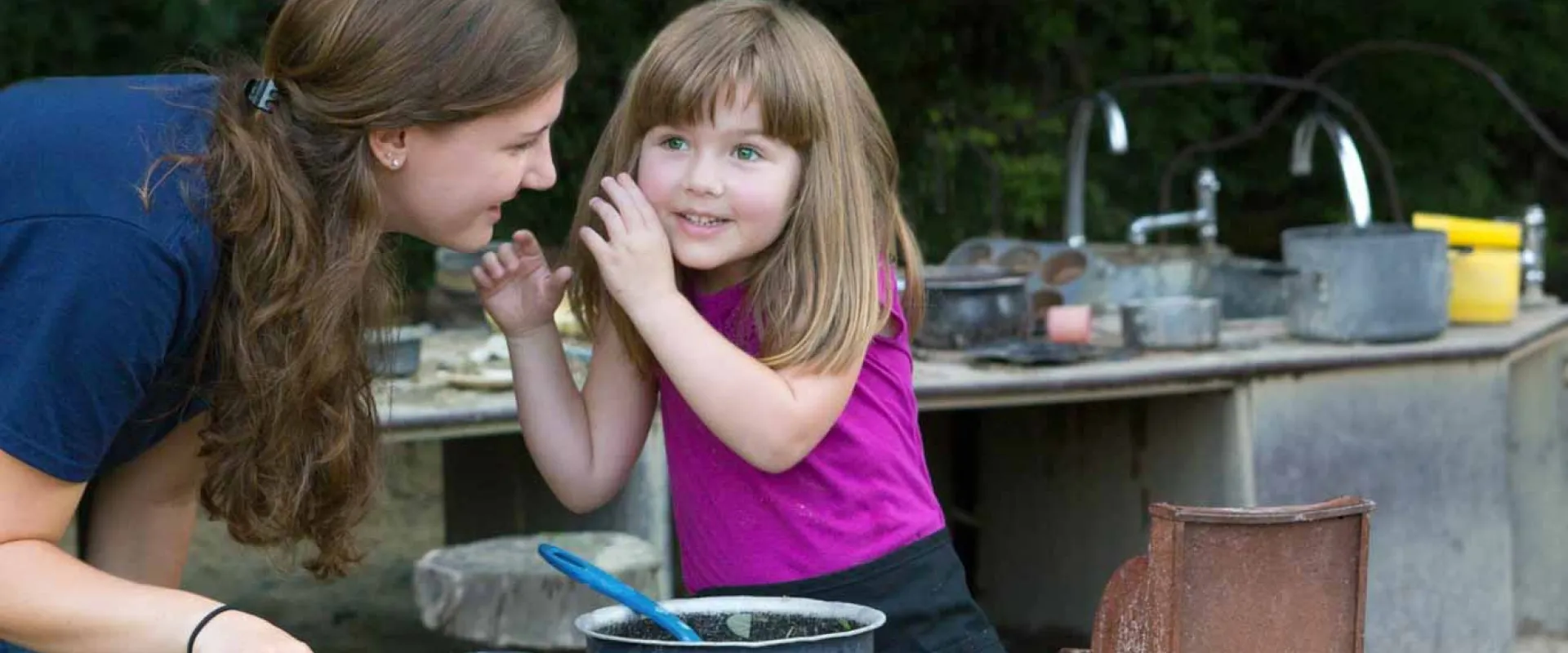 An Educator is talking with a young white girl in pink at the Mud Cafe with a steel counter behind her that is littered with kitchen utensils.