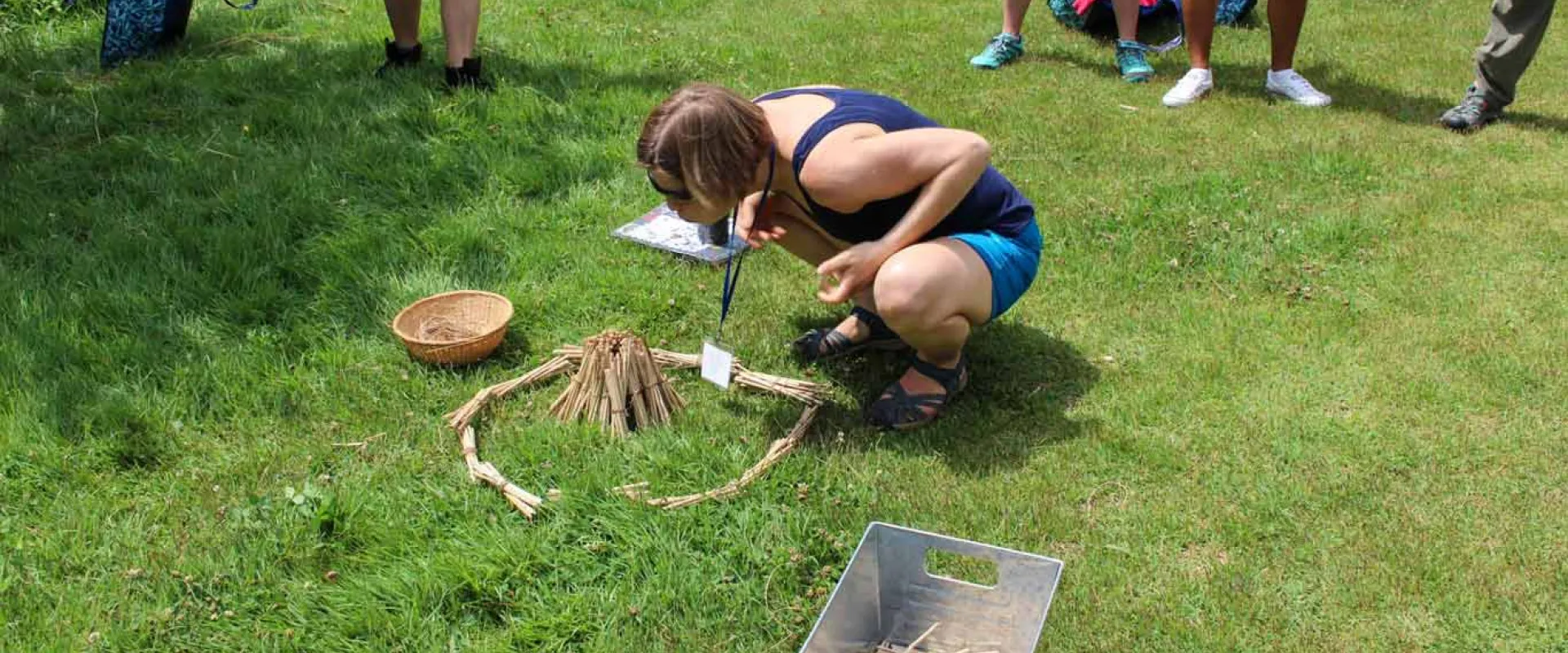 A woman with short hair squats on the grass, leaning over a pile of sticks surrounded by a ring of sticks, resembling a fire pit. The feet of four other peoples feet, standing around her observing can be seen in the background.  A bowl and metal bin lay in the grass next to her.