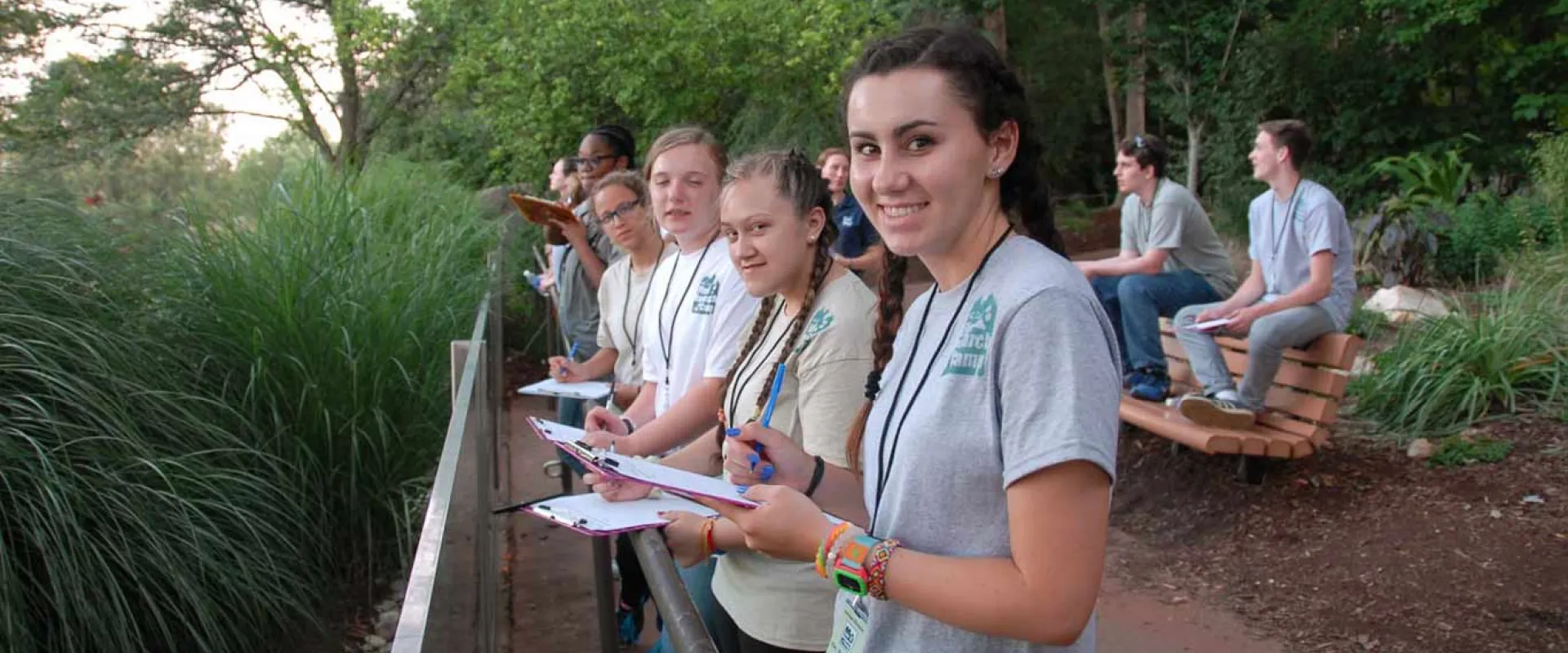 Seven students standing on a walkway outdoors, looking over a railing. The students are wearing matching gray or white t-shirts, some with lanyards, holding clipboards and pencils, appearing to be taking notes. The young woman in the foreground is smiling at the viewer. Tall, dense green grasses are on the left, and a few other students are seated on a wooden bench on the right, with a dirt path in front of them. The background is a mix of trees and foliage.