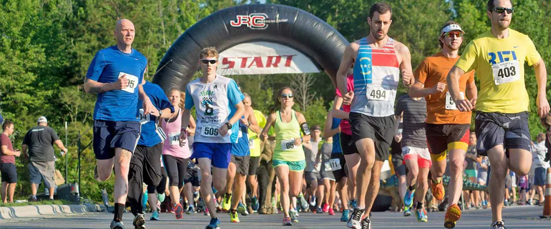 A large group of runners is starting a road race, moving forward beneath a large black inflatable arch labeled "START". The runners are wearing various brightly colored athletic gear.