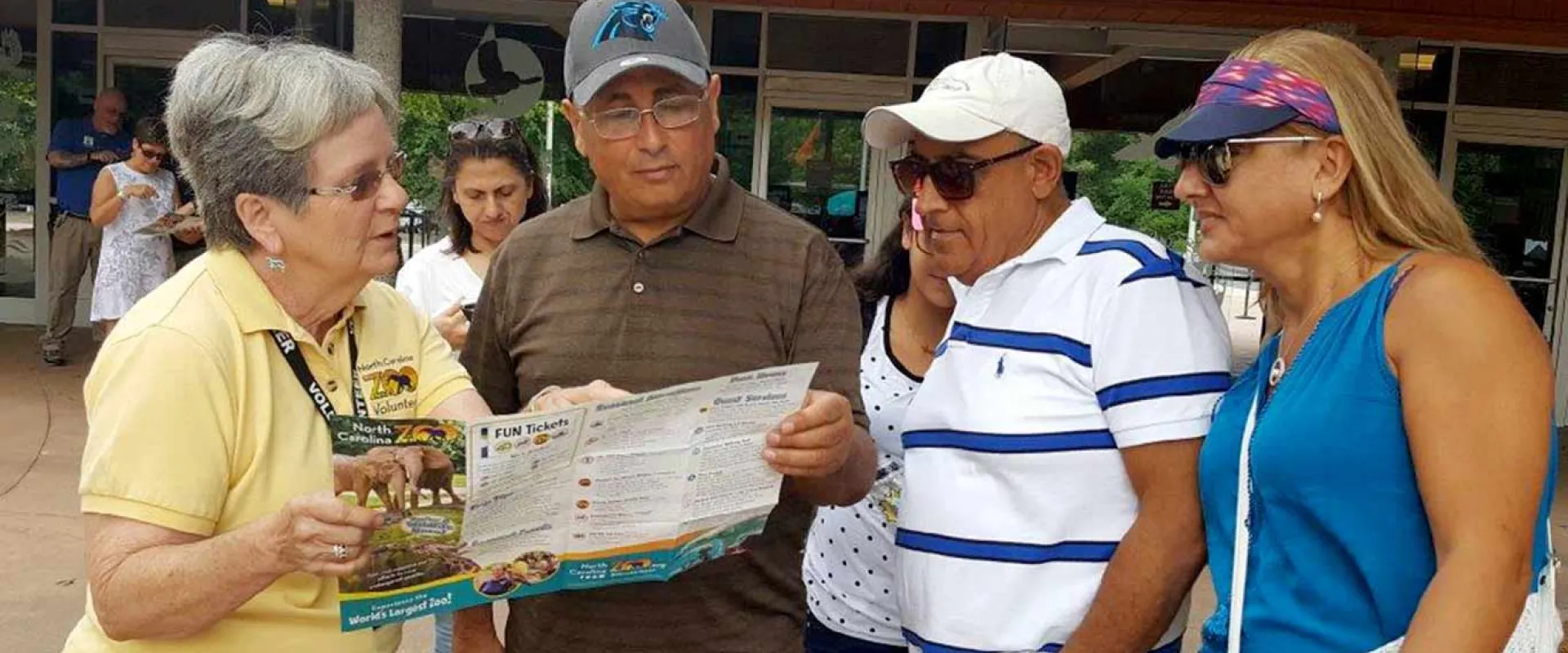 A zoo or park employee in a yellow shirt is helping three visitors by pointing to information on a folded map or brochure. They are standing outside near the entrance to a building.