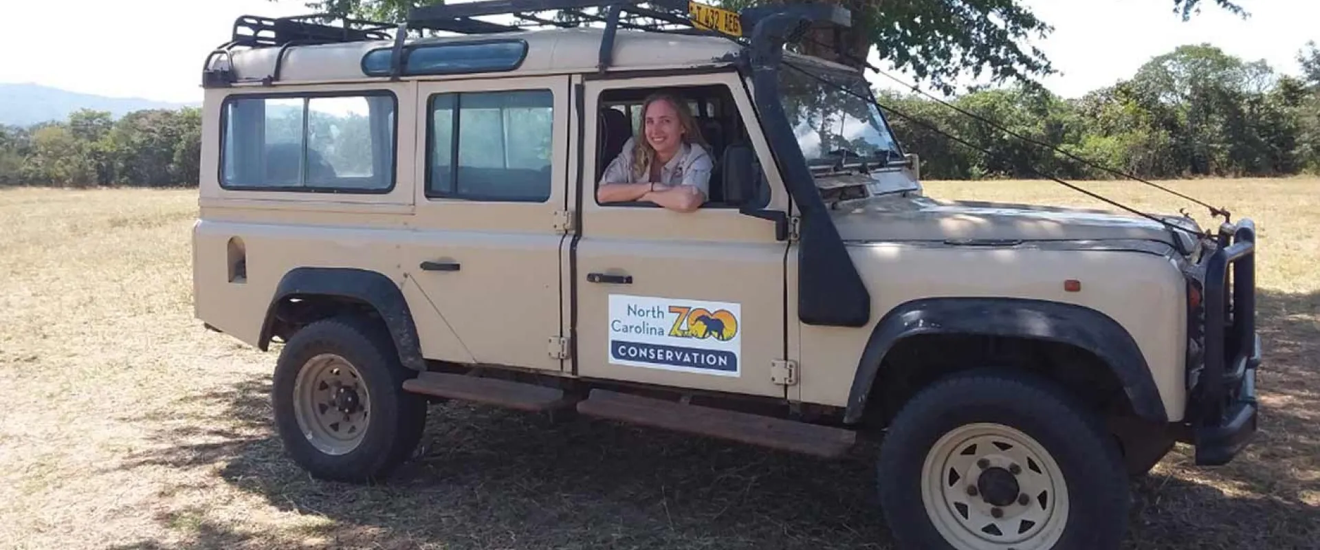 A light tan Land Rover Defender or similar safari-style off-road vehicle is parked in a sunny, dry field with trees in the background. A person with light hair is visible leaning out of the open driver's side door.