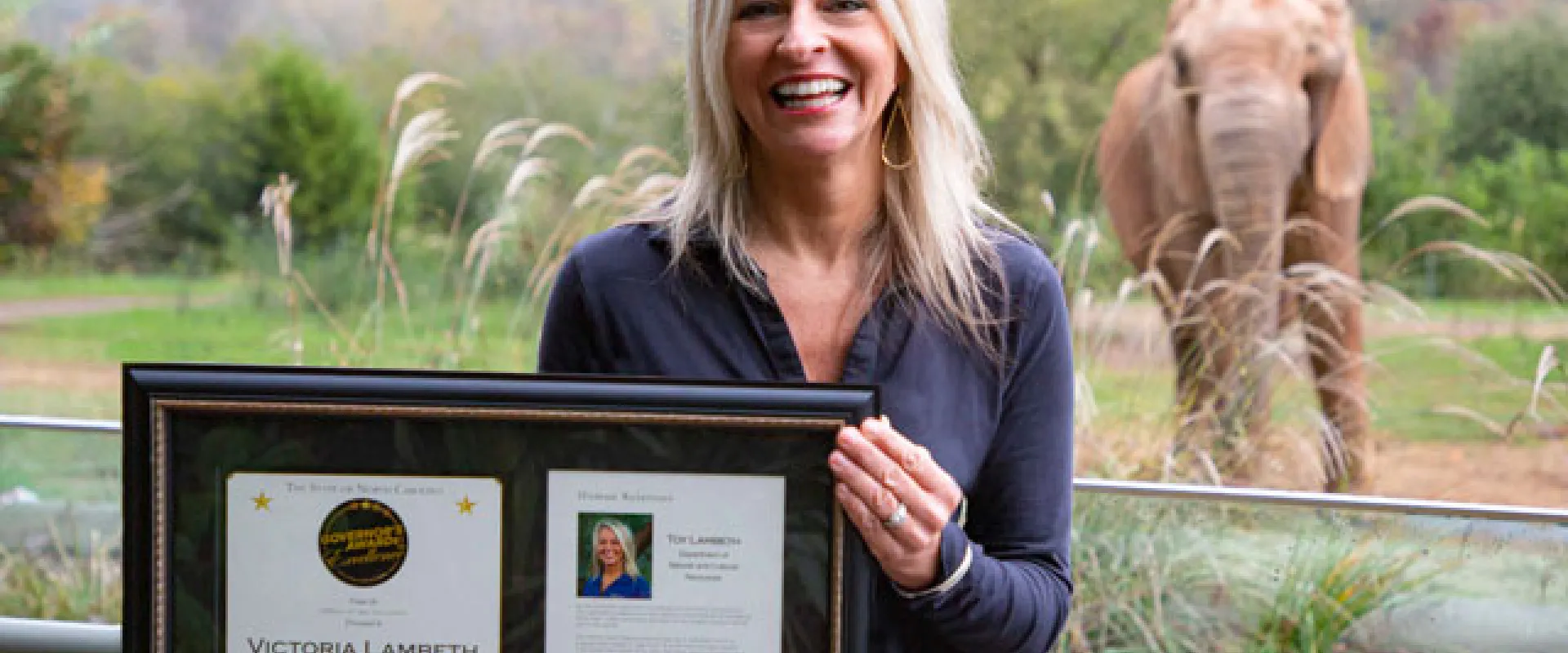 A smiling woman with long blonde hair, wearing a dark blue shirt, stands outdoors holding a framed document or award with two sections. In the background, behind a low metal railing, a young, reddish-brown elephant is visible standing in a grassy enclosure.