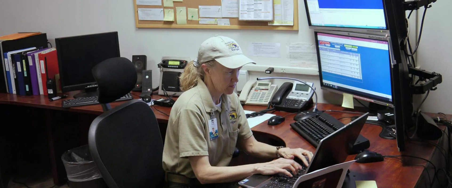 An adult man wearing a tan uniform shirt and a white baseball cap is seated at a desk in an office, working on a computer with multiple monitors.