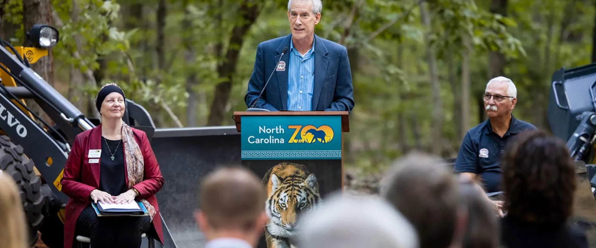 A man in a blue jacket is speaking at a podium with the North Carolina Zoo logo on the front during an outdoor press conference. Two other people are seated beside him, with construction equipment visible in the background.