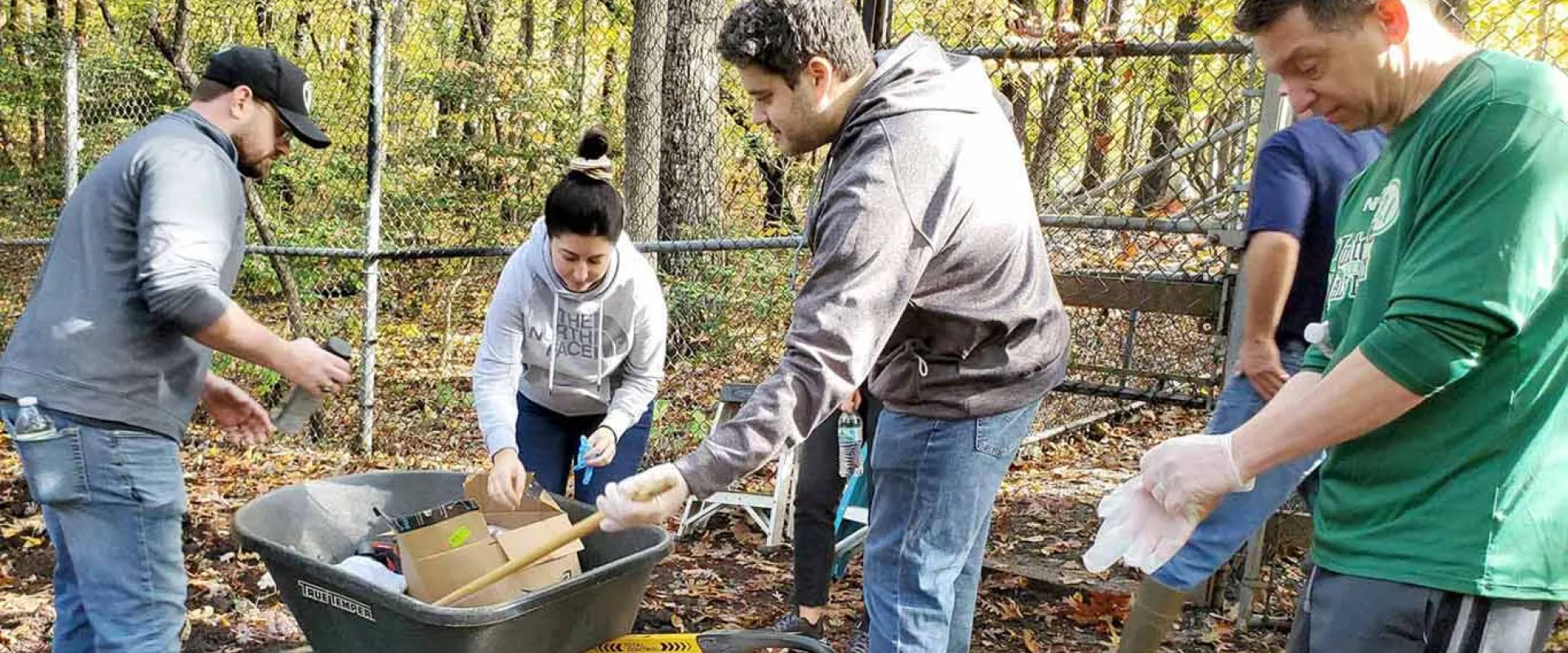 A group of five volunteers is working outdoors in a wooded area surrounded by a chain-link fence, loading various items, including cardboard, into a wheelbarrow.