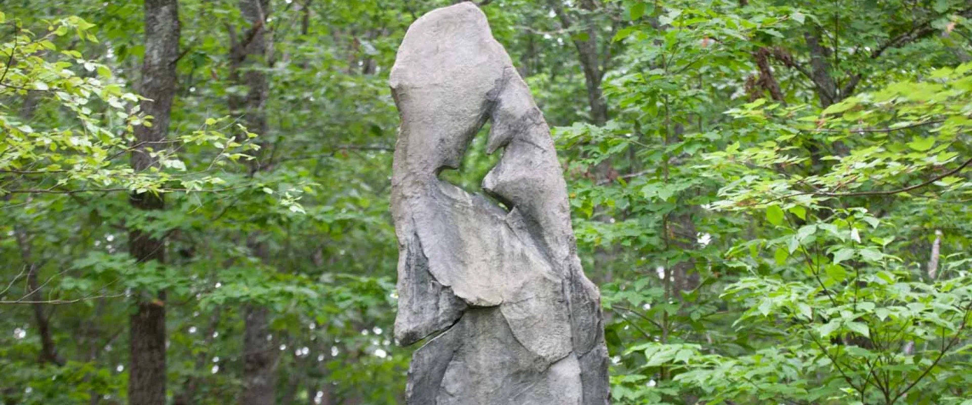 A gray, vertical, abstract outdoor sculpture, possibly made of carved stone or concrete, featuring smooth curves and a large negative space shaped like a bird in flight. The sculpture is positioned against a backdrop of dense, leafy green trees.