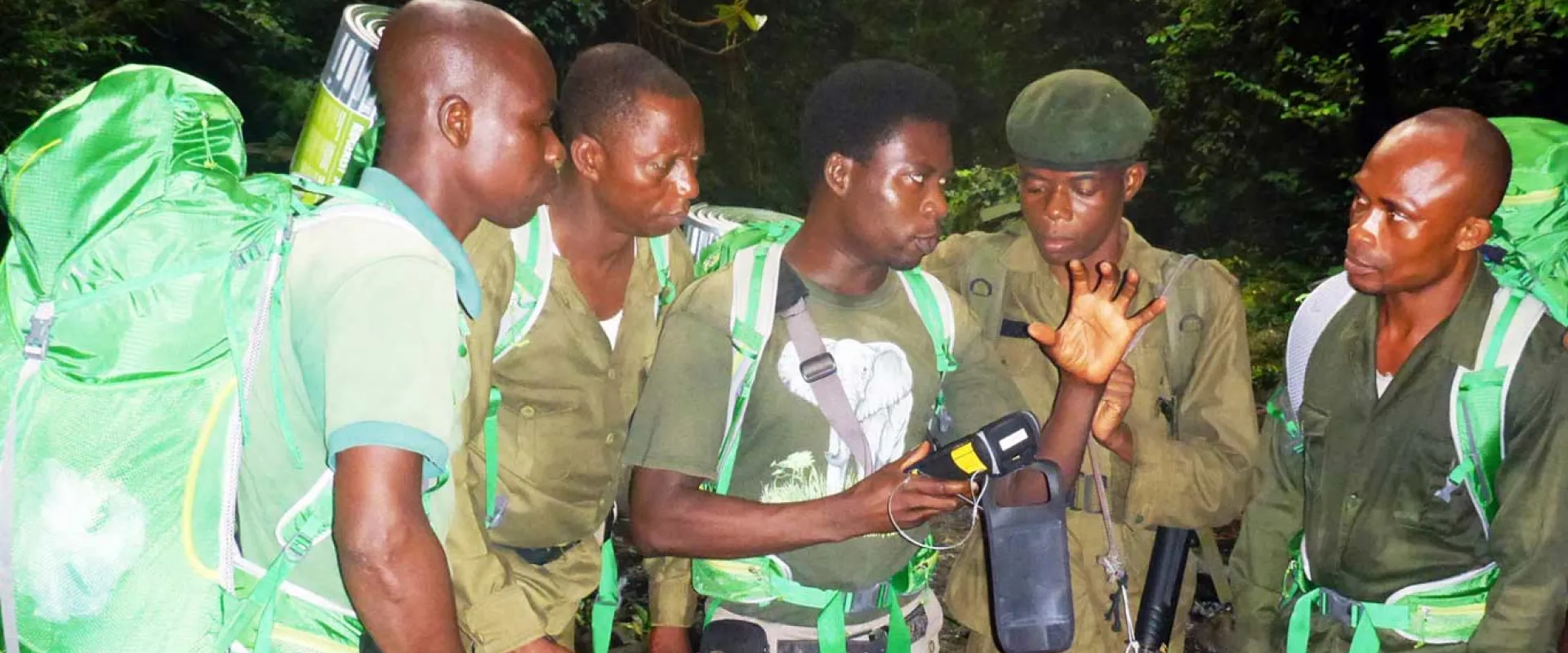 Five men, wearing matching short-sleeved green ranger uniforms and shoulder bags, stand close together in a dense forest or jungle environment. They are looking intently at a small electronic device, likely a smartphone or GPS unit, held by the man on the left. The ground beneath them is a dirt path, and lush green foliage surrounds them.