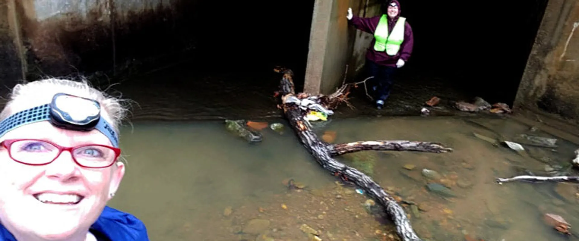 A low-angle selfie of two women standing in front of a dark concrete storm drain outlet. The woman closest to the camera is wearing a blue jacket, red glasses, and a headlamp, and is smiling. The second woman stands in the background, inside the dark entrance, wearing a dark jacket and a high-visibility safety vest. The ground  is a shallow stream of muddy water containing rocks, trash, and a large piece of driftwood. 