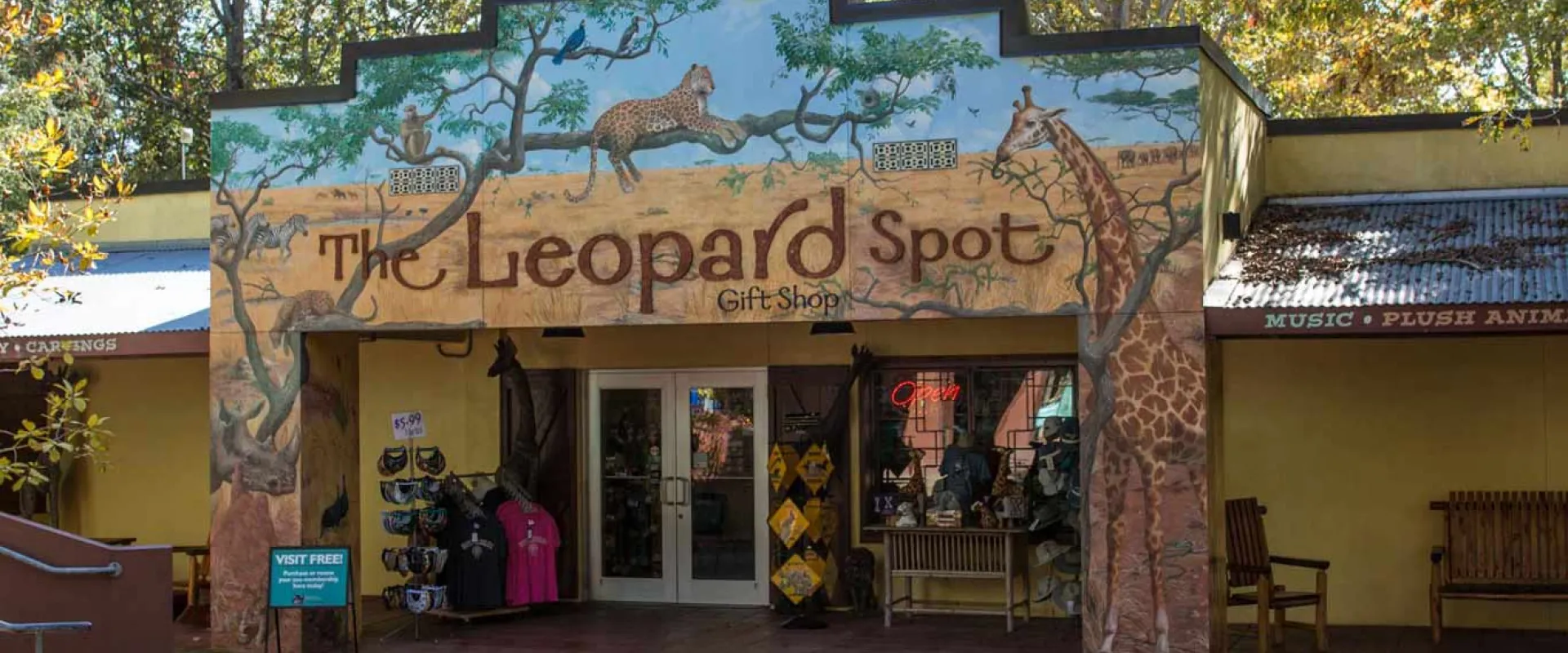 The front of a small tan building, possibly a gift shop. On the brown roof is a sign with the image of an African savanna, a giraffe and a leopard lounging in a tree that reads, "The Leopard Spot." Trees line the back of the building."