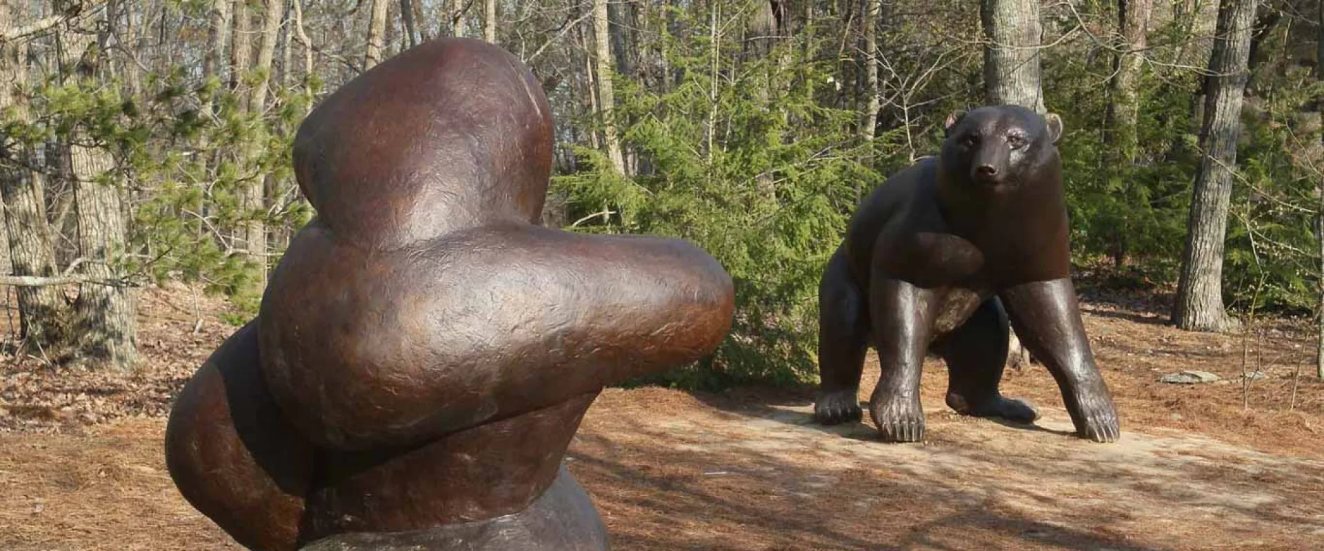 Two large bronze sculptures are displayed outdoors in a sunny, forested area. In the foreground is a smooth, abstract, dark brown sculpture, and in the background is a realistic bronze sculpture of a small bear standing on all fours.