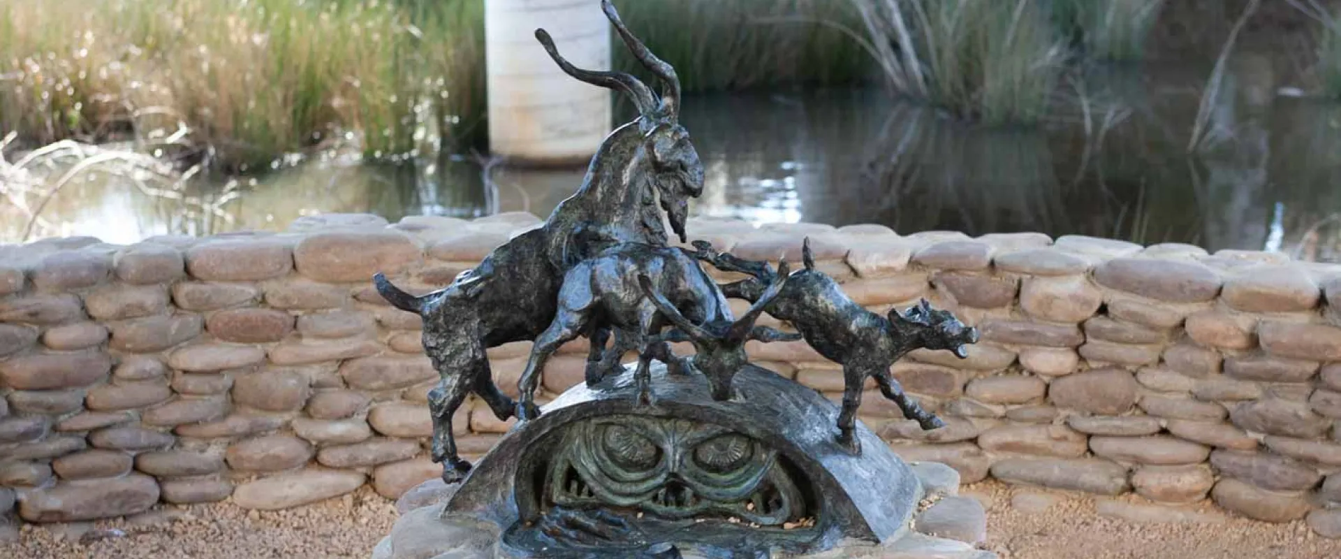 A bronze sculpture of three goats, with swirling forms and intricate details, atop a circular pedestal of stacked, light-colored stones. In the background, a calm body of water and two concrete pillars of a bridge.