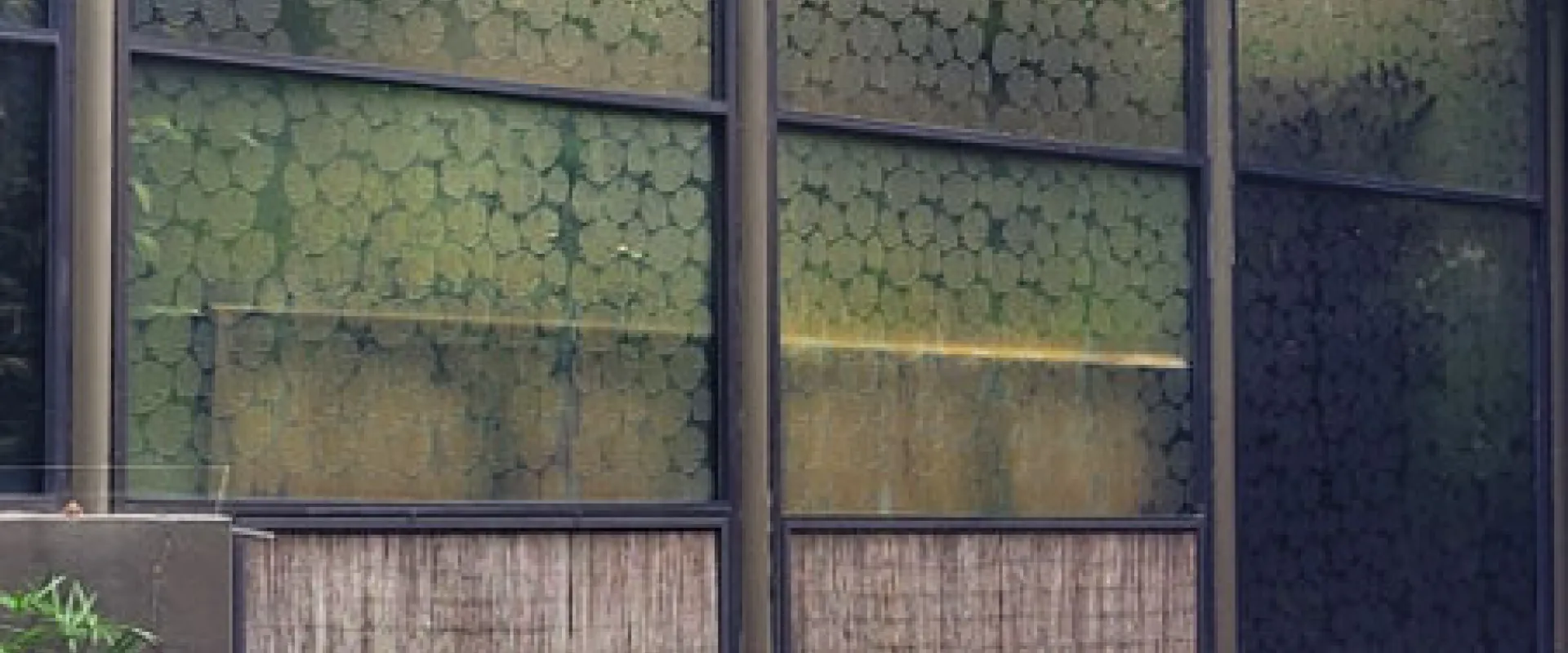  A long, glass-paneled building with a white fabric roof is shown with several flamingos standing in a small pool of water in the foreground. The glass panels have a translucent leaf pattern on them, obscuring the view into the building. T