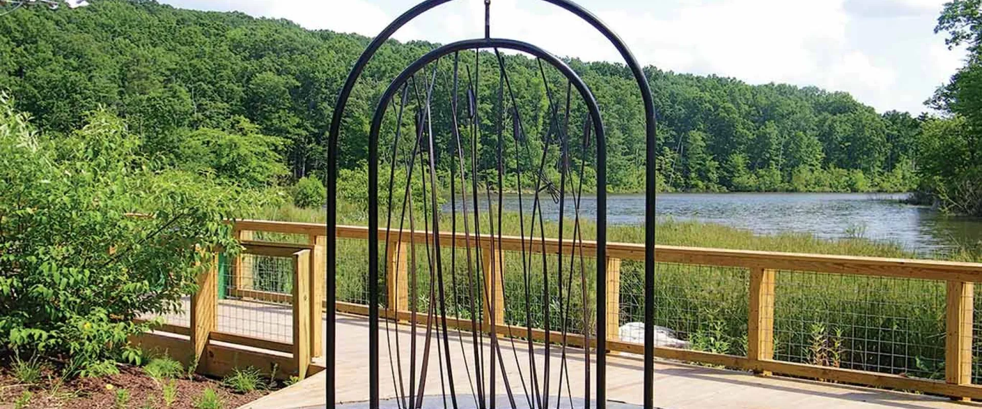 A tall, black metal outdoor art installation made of vertical rods framed by two large arches. The sculpture is centered on a paved area next to a wooden boardwalk or deck that overlooks a calm lake or pond. The area surrounding the lake is covered in green reeds, and a dense, lush green forest covers the rolling hills in the background under a bright, partly cloudy sky.
