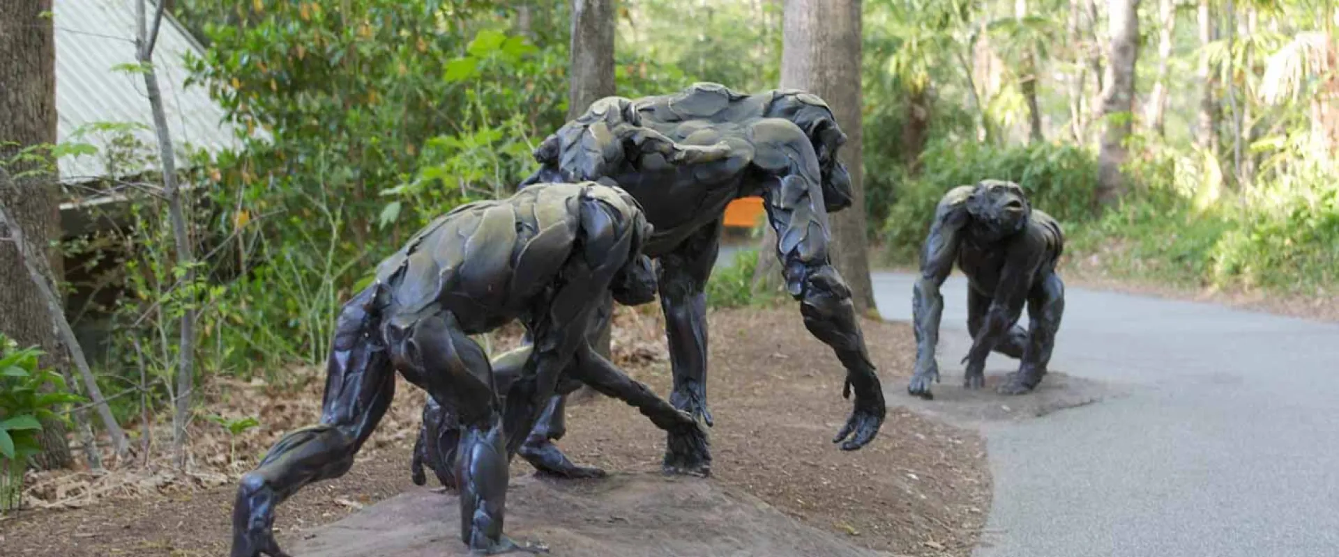 A bronze sculpture of a family of three chimpanzees stands on a raised patch of dirt next to a paved path. The sculpture features a large male gorilla standing over a female and a younger gorilla. The sculptures have a textured, segmented look, as if made from individual plates of metal. The path curves away to the right, and the area is surrounded by trees and green plants.