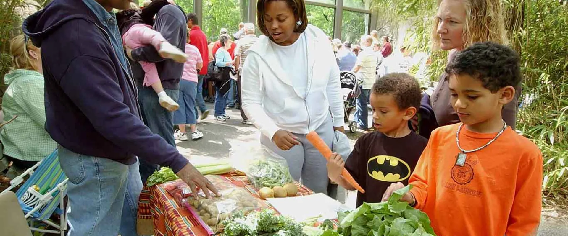 A man wearing glasses, jeans, and a NC Zoo uniform stands behind a table laden with fresh produce next to an animal habitat in the woods, gesturing towards the leafy greens. A woman, possibly a mother, stands next to him, and two young boys, possibly her children, look at the vegetables. One boy wears a black t-shirt with a yellow Batman logo, and the other wears an orange t-shirt. Other guests are visible in the background, surrounded by greenery.