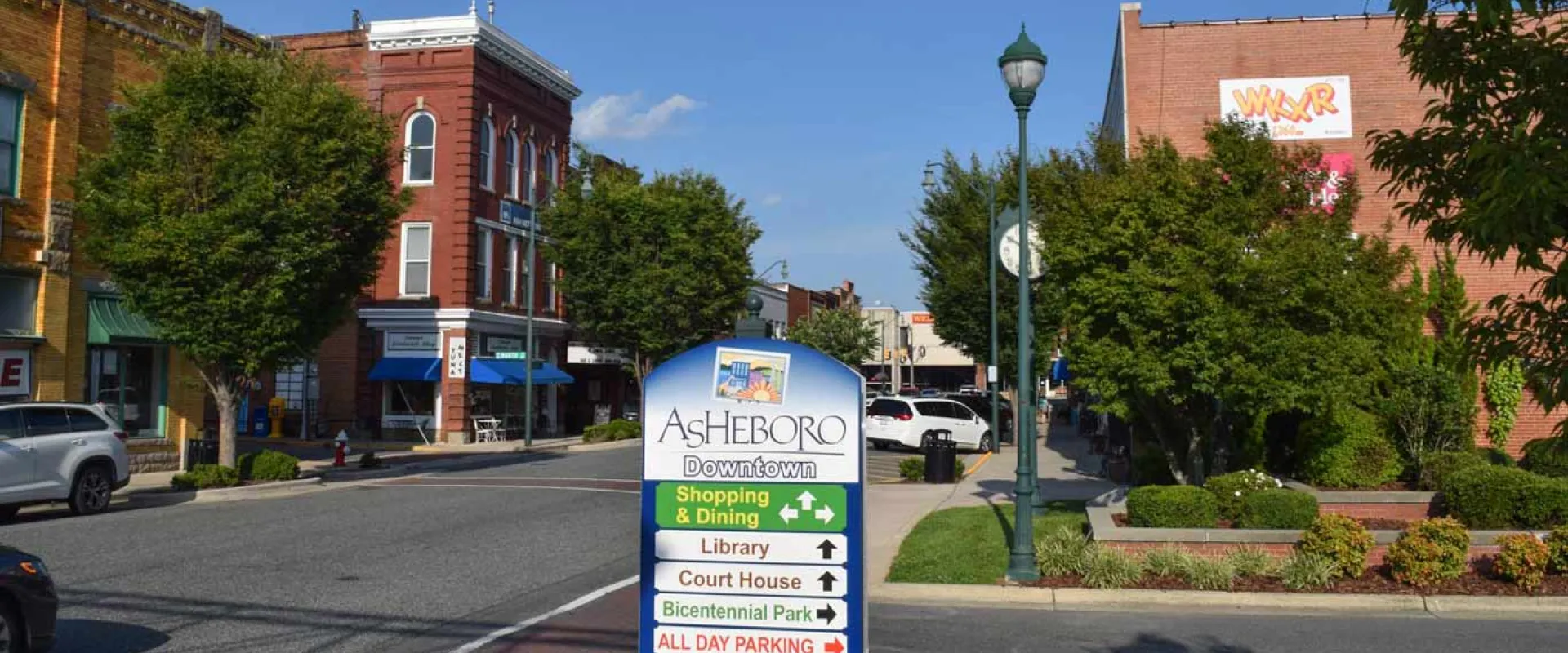 An informational sign with a blue base stands in the middle of a sunlit, tree-lined downtown street in front of two and three-story brick buildings.