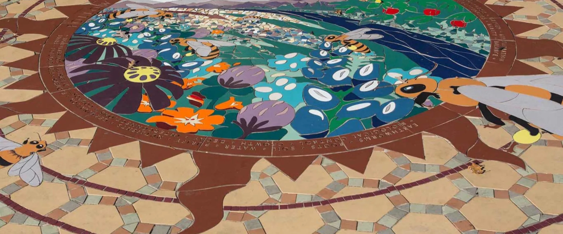 Follow the Pollen Path, a ceramic tile mural, inlaid in front of the Garden Friends playground. This circular mural has an orange sun shape inside with a colorful outdoor scene in the center.