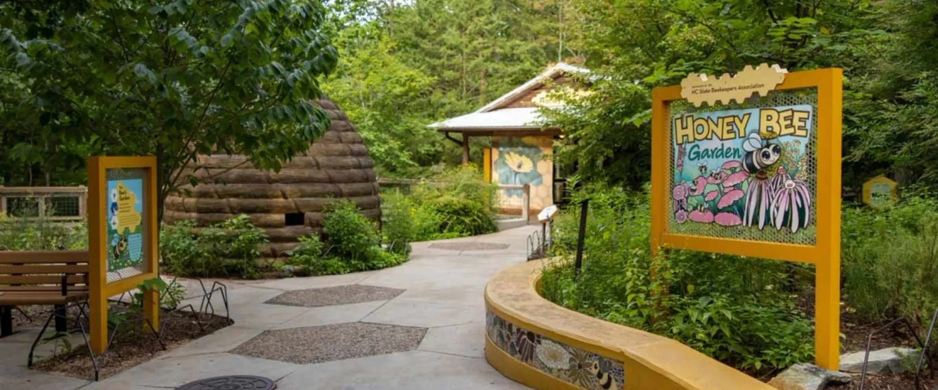 A concrete pathway winds through a lush, green outdoor exhibit. On the right, a large yellow sign reads "HONEY BEE GARDEN" in yellow and black text, featuring an illustration of a Honey Bee. A low, curved yellow wall runs alongside the path, and behind it, dense foliage. To the left, another yellow sign stands near a wooden bench. Further down the path, a rustic log beehive structure is visible among the trees. 