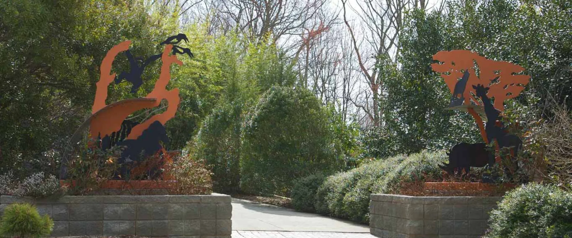 A panoramic view of the entrance to an outdoor path flanked by two large, metal sculptures of animals in dark brown or black against a rusted orange background, depicting scenes like a giraffe eating from a tree on the right, and other African animals, possibly including a lion, on the left. The path is brick and concrete and leads between two low stone or concrete planter walls topped with shrubbery.