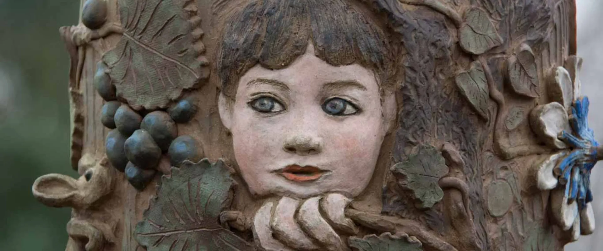 A close-up of a detail on a large, highly decorated totem pole that features a brightly colored wooden carving on a post, featuring a child's face surrounded by grape leaves and bunches of purple grapes