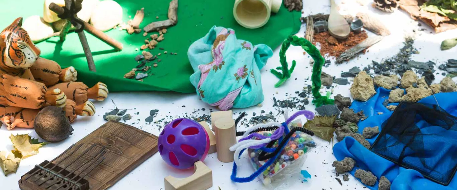 A table covered in a white cloth and littered with a variety of craft items including a toy tiger, green clay, fuzzy pipecleaners, pinecones, green paper, sand, sticks, wooden blocks, and a blue plastic material.