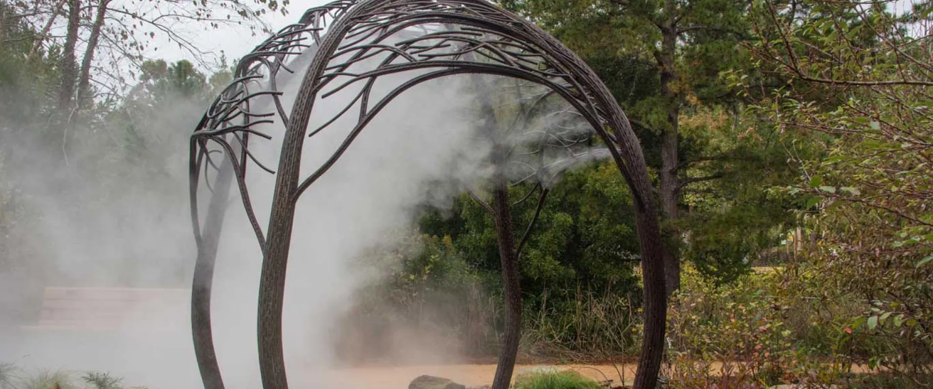 A decorative arch that is shaped to look like the bent branches of trees that spreads across a walkway that is surrounded by trees and bushes. A soft cloud of misting water sprays from the middle of the archway.