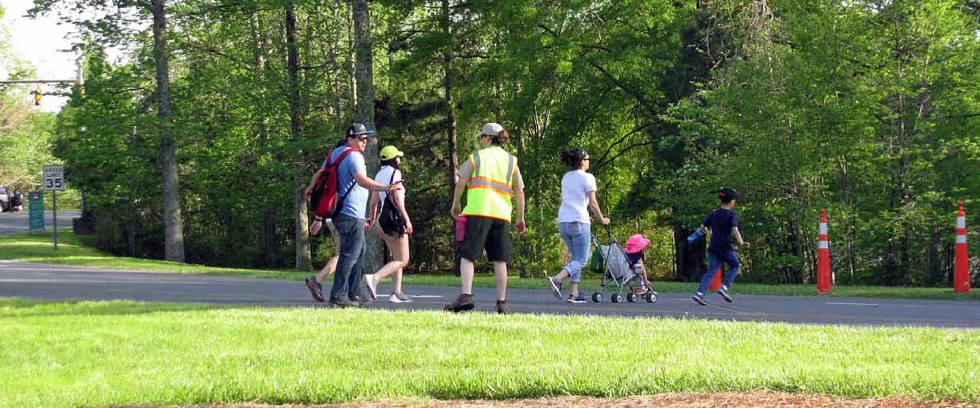 A diverse group of people is walking along a paved path, with a large, grassy area to their left and trees and bushes to their right. The group includes two adults pushing a stroller, a couple, and a man in a bright yellow safety vest who appears to be directing or monitoring the group.