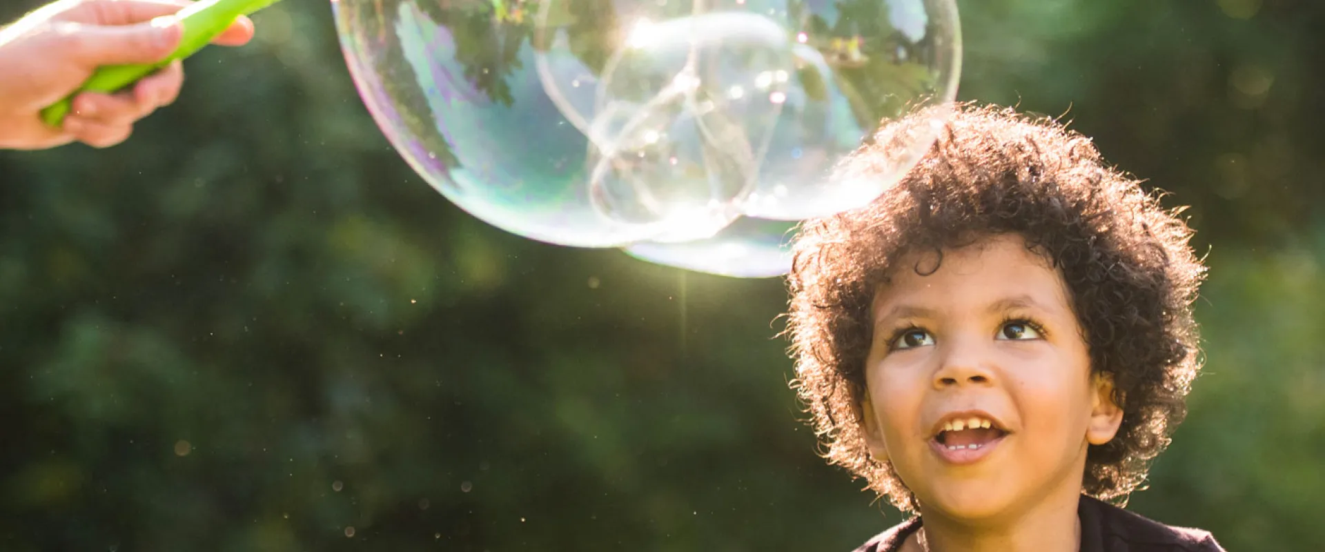 A child with short, curly hair enjoying some time with giant bubbles outside in the sun.