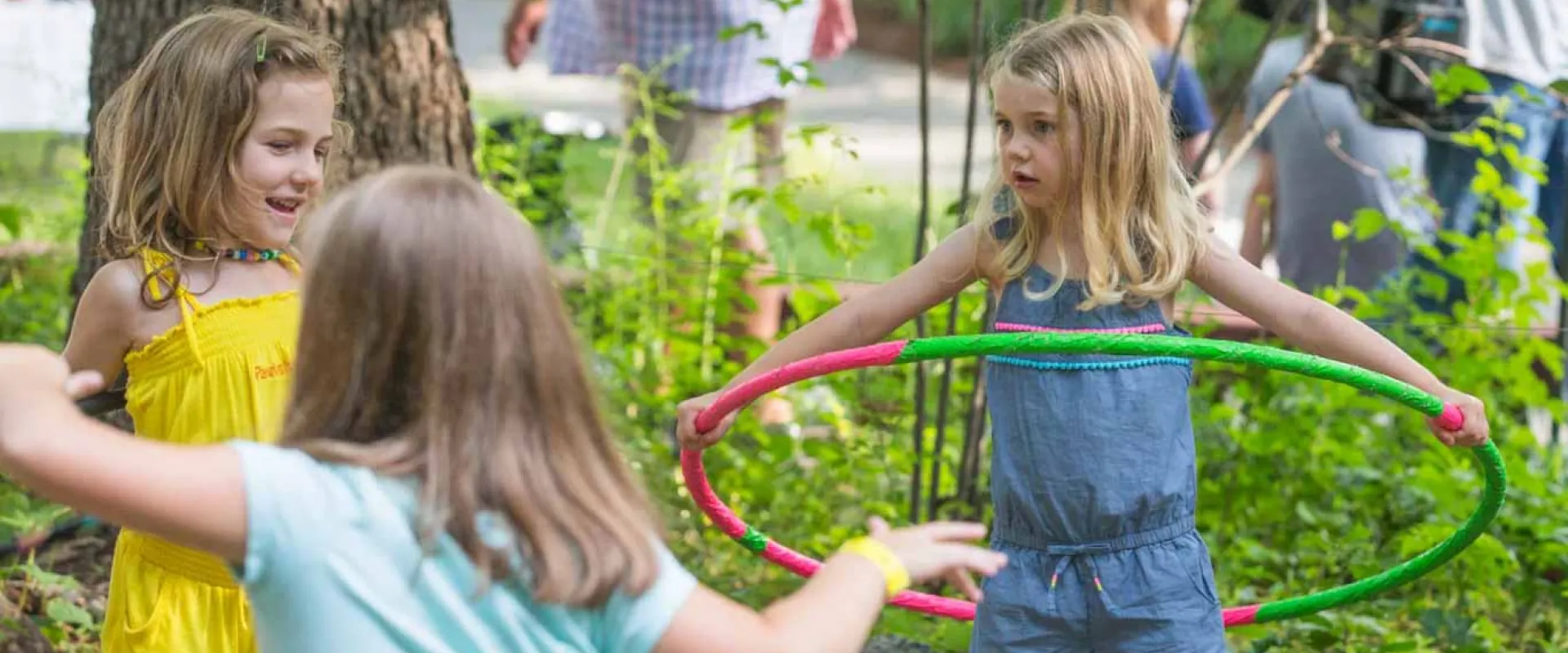 Three young girls playing in a lush garden area. The girl on the far right has a red and green hula hoop around her waist, while the other two girls appear to be running around. Adults can be seen in the background walking about.