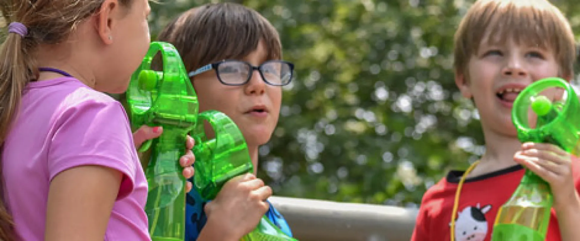 A group of young campers holding green personal fans stand together, posing in front of a large brass statue of a Bison in a wooded area.