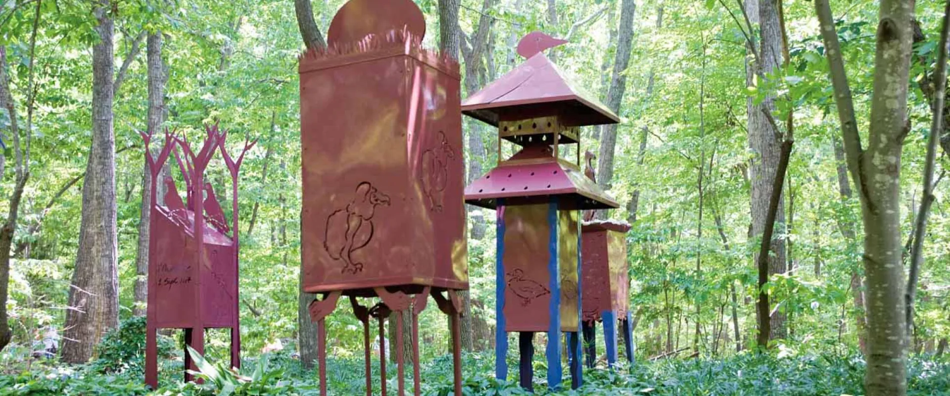 A group of tall, artistic metal sculptures resembling red-brown lanterns or towers, featuring animal cut-outs, are displayed outdoors in a dense wooded area.