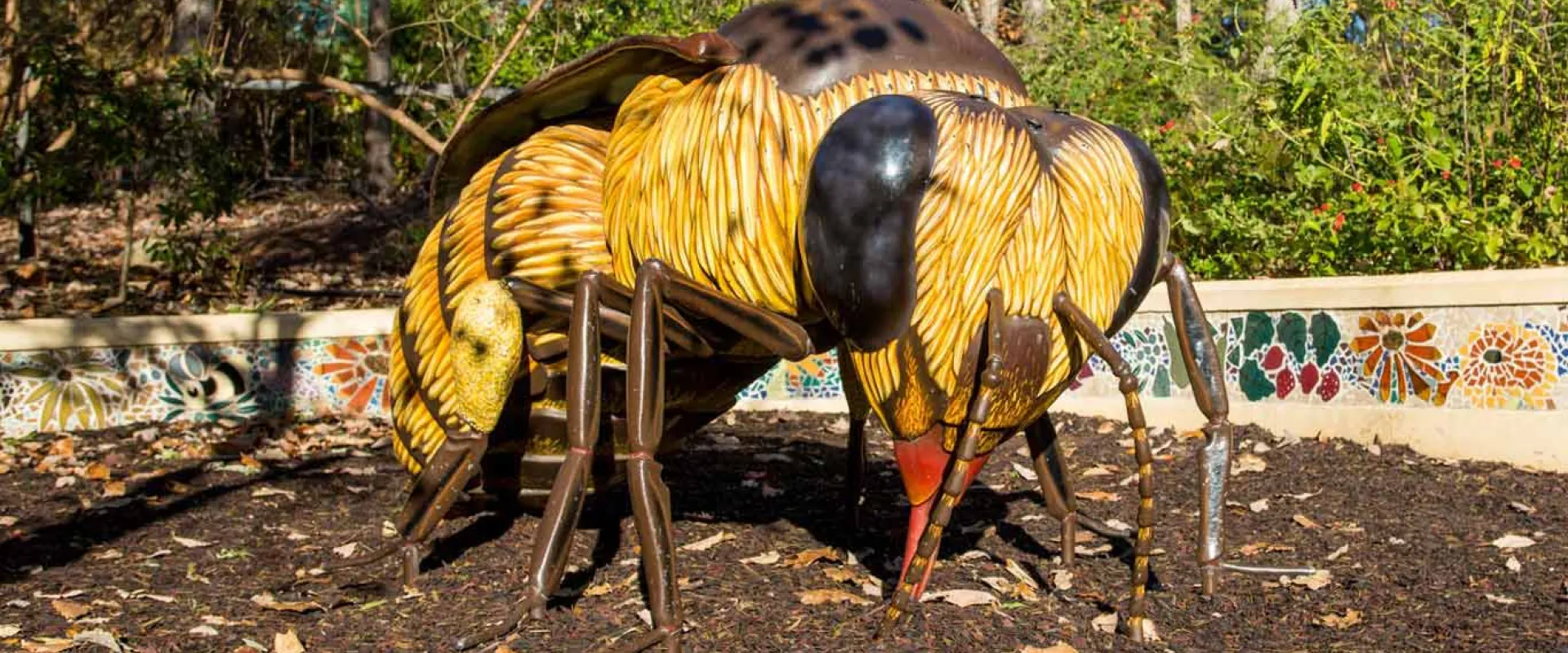 A gigantic statue of a worker bee stands on the ground at the Garden Friends Playground with its wings tucked back. A mosaic wall stands behind it with tiled words that read "Bee Kind" and various small flowers.