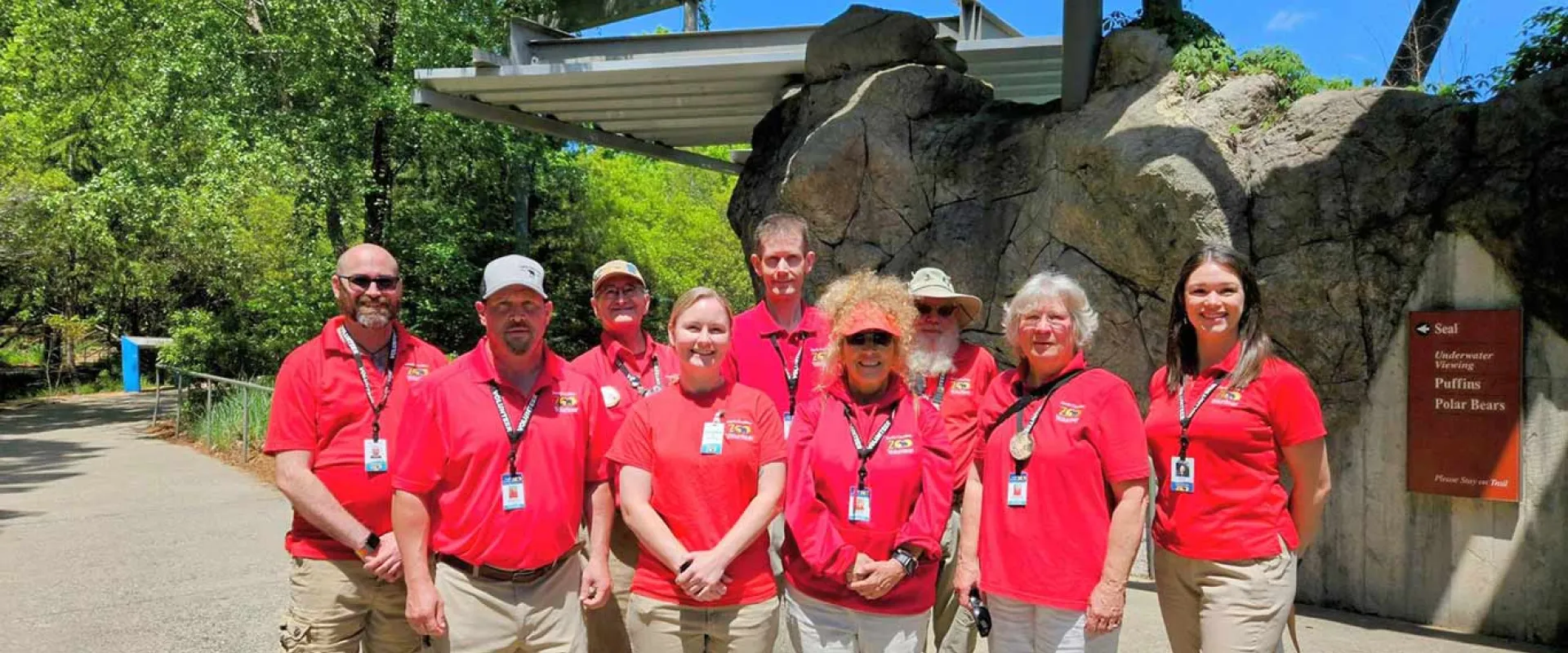 Group of nine Zoo volunteers standing in front of a large rock walk next to trees