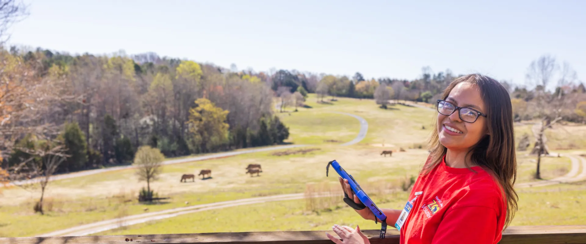 Research volunteer in a red shirt smiles at camera while overlooking the Watani grasslands habitat. Rhinos and antelope are on the hills in the background on a bright sunny day.
