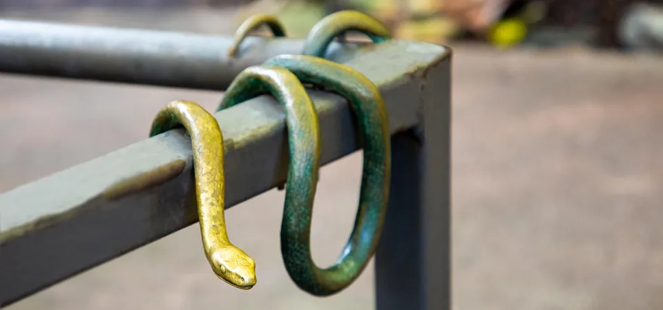 A life-like bronze sculpture of a snake draped on and around a brown metal handrail at the North Carolina Zoo.