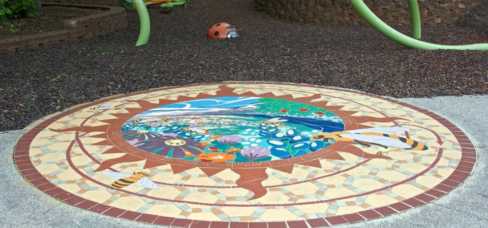 Follow the Pollen Path, a ceramic tile mural, inlaid in front of the Garden Friends playground. This circular mural has an orange sun shape inside with a colorful outdoor scene in the center. 